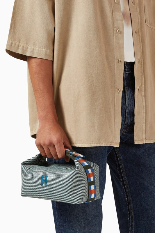 Person holding a Hermès Small Bride-à-Brac au Carré Toiletry Bag Green Forest H103802Mv03 with a brand logo, wearing a beige shirt and blue jeans.