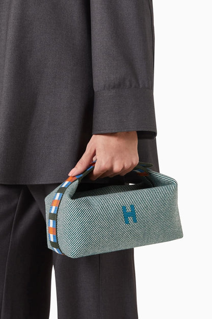 Person holding a Hermès Small Bride-à-Brac au Carré Toiletry Bag Green Forest H103802Mv03 with a logo, wearing a dark gray coat and pants.