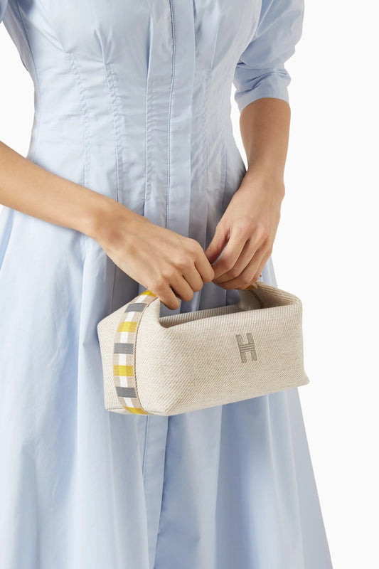 Person holding a Hermès Small Bride-à-Brac au Carré Toiletry Bag Seige H103802Mv01 bag with a brand logo, wearing a light blue dress.