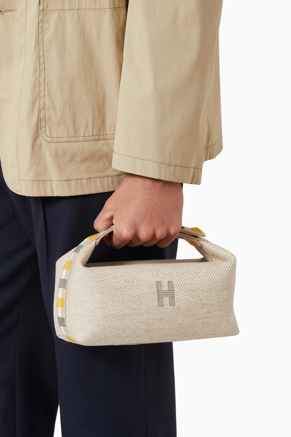 Person holding a Hermès Small Bride-à-Brac au Carré Toiletry Bag Seige H103802Mv01 with a logo, wearing a beige jacket and dark pants.