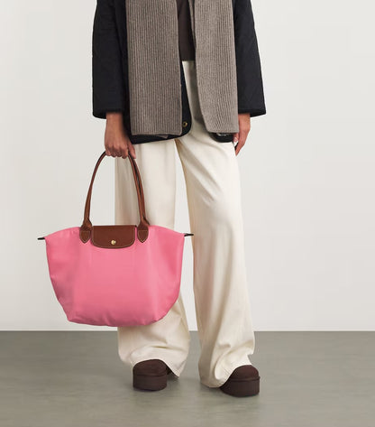 Person holding a Le Pliage Original L Tote Bag Marshmallow L1899089P92 with brown handles and trim, wearing a black coat, beige scarf, white pants, and brown shoes.