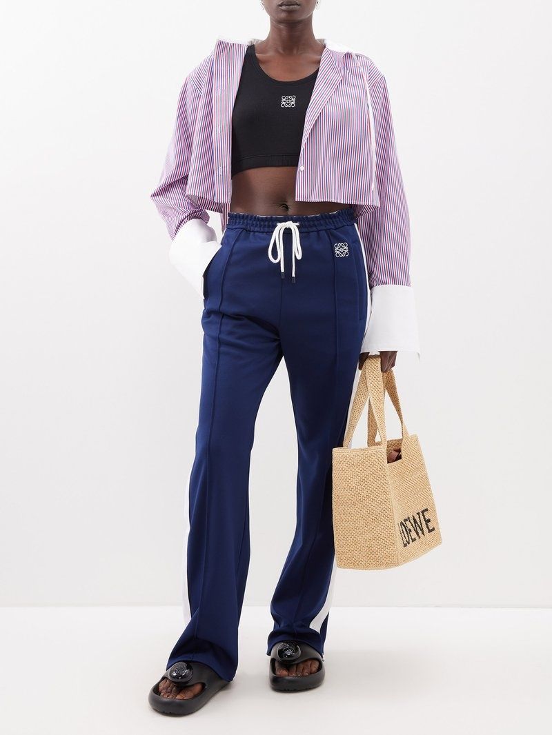 Person wearing a purple cropped hoodie, black top, and blue pants, holding a straw bag with Loewe Paula Ibiza Foam Thong Slide Black L616S03X02 1100.