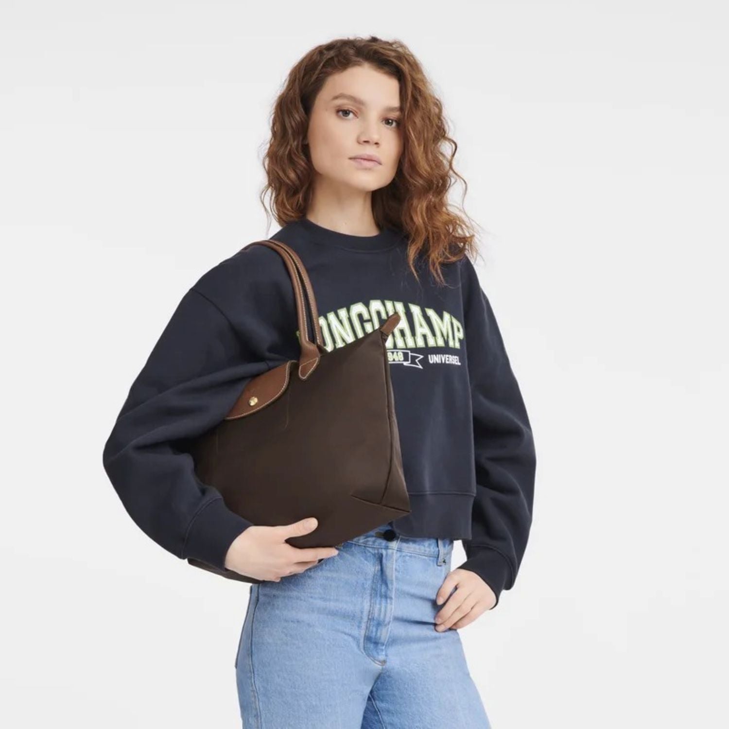 Woman wearing a navy sweatshirt with 'Longchamp' branding, holding a Longchamp Le Pliage Original M Tote Bag Ebony L2605089P81 against a white background.