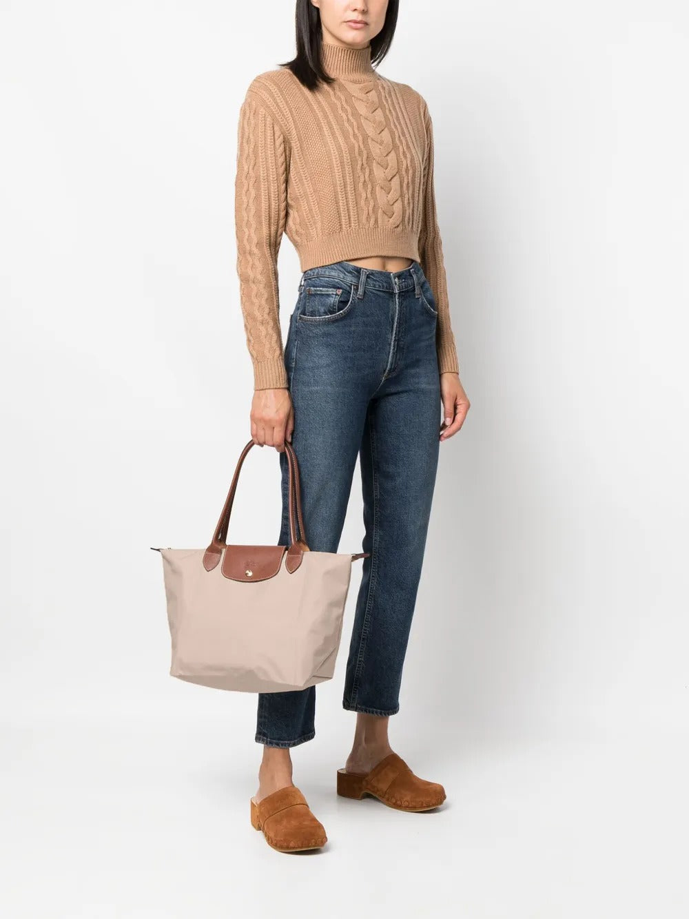 Woman holding a Longchamp Le Pliage Original M Tote Bag – Pebble L2605089349 wearing a beige sweater, blue jeans, and brown shoes on a white background