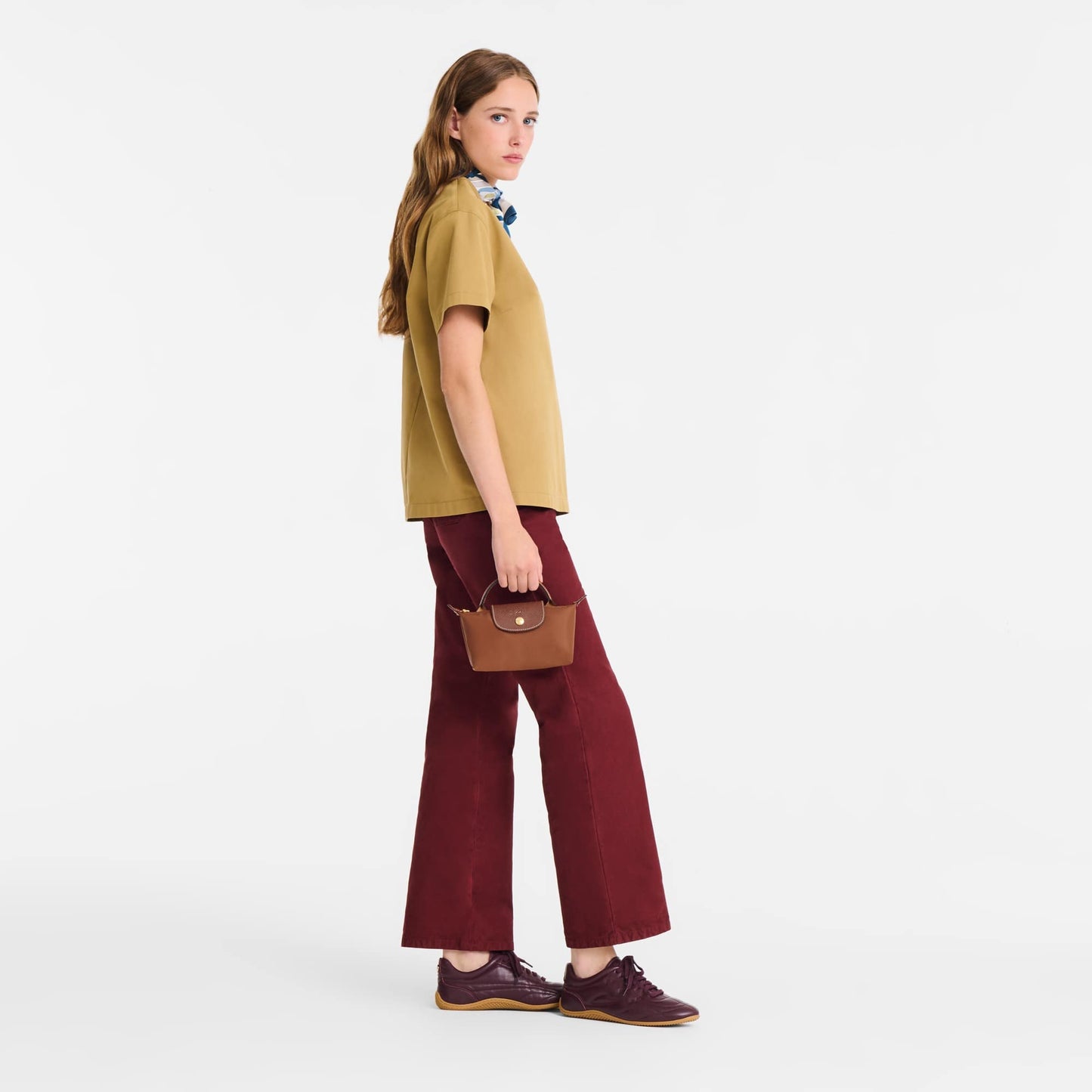 Woman wearing a mustard shirt and red pants holding a Longchamp Le Pliage Original Pouch with Handle – Cognac 34175089504 on a white background