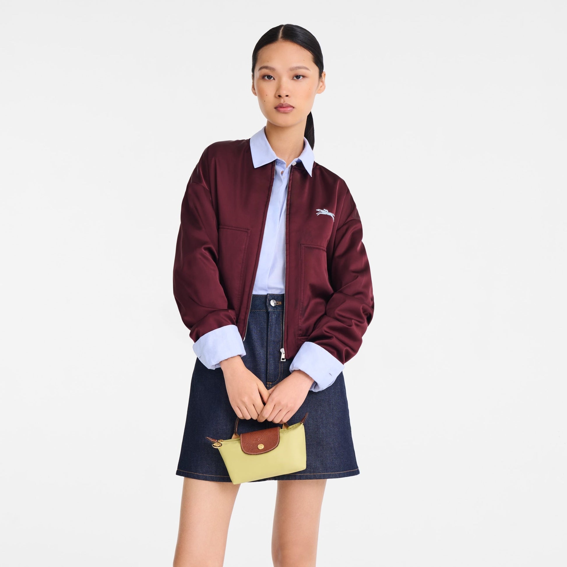 Person wearing a maroon jacket with a brand logo, white shirt, and denim skirt holding a yLongchamp Le Pliage Original Pouch with Handle – Pistachio 34175089244 on a white background