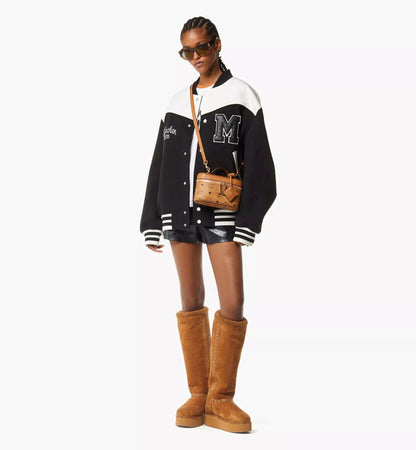 Person wearing a black and white varsity jacket with a letter 'M', shorts, and brown knee-high boots holding MCM Aren Vanity Case in Visetos Leather Mix Cognac MWRGSTA02CO001 on a white background.