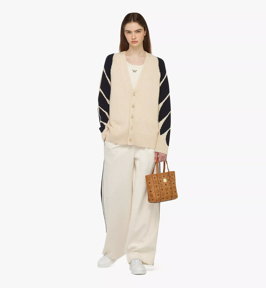 Woman wearing a beige vest, black and white striped cardigan, and white pants, holding a MCM Mini Toni Top-Zip Shopper in Visetos Cognac MWPAATN04CO001 on a white background.