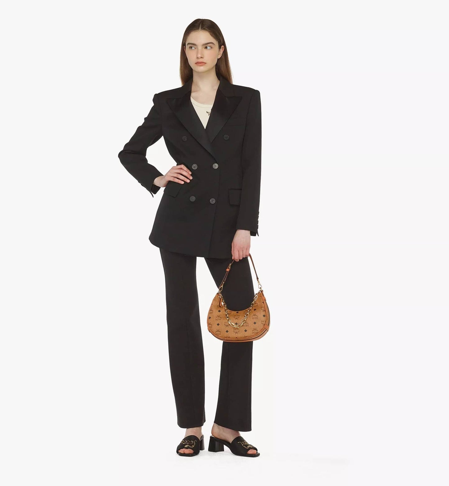 Woman wearing a black suit holding a MCM Small Aren Crescent Hobo Bag in Visetos Cognac MWHCATA11CO001 on a white background