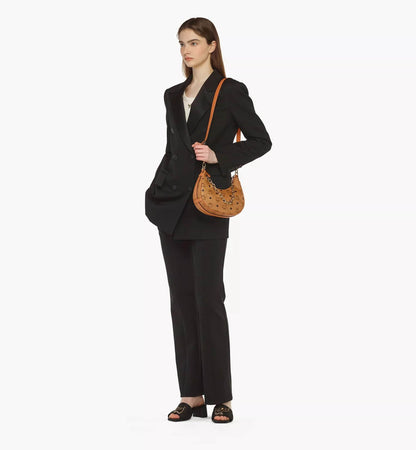 Woman in a black suit holding an MCM Small Aren Crescent Hobo Bag in Visetos Cognac MWHCATA11CO001 on a white background