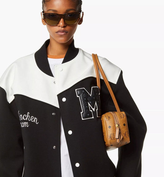 Person wearing a black and white varsity jacket with a 'M' emblem, holding a MCM Small Aren East West Shoulder Bag in Visetos Cognac MWSGSTA02CO001