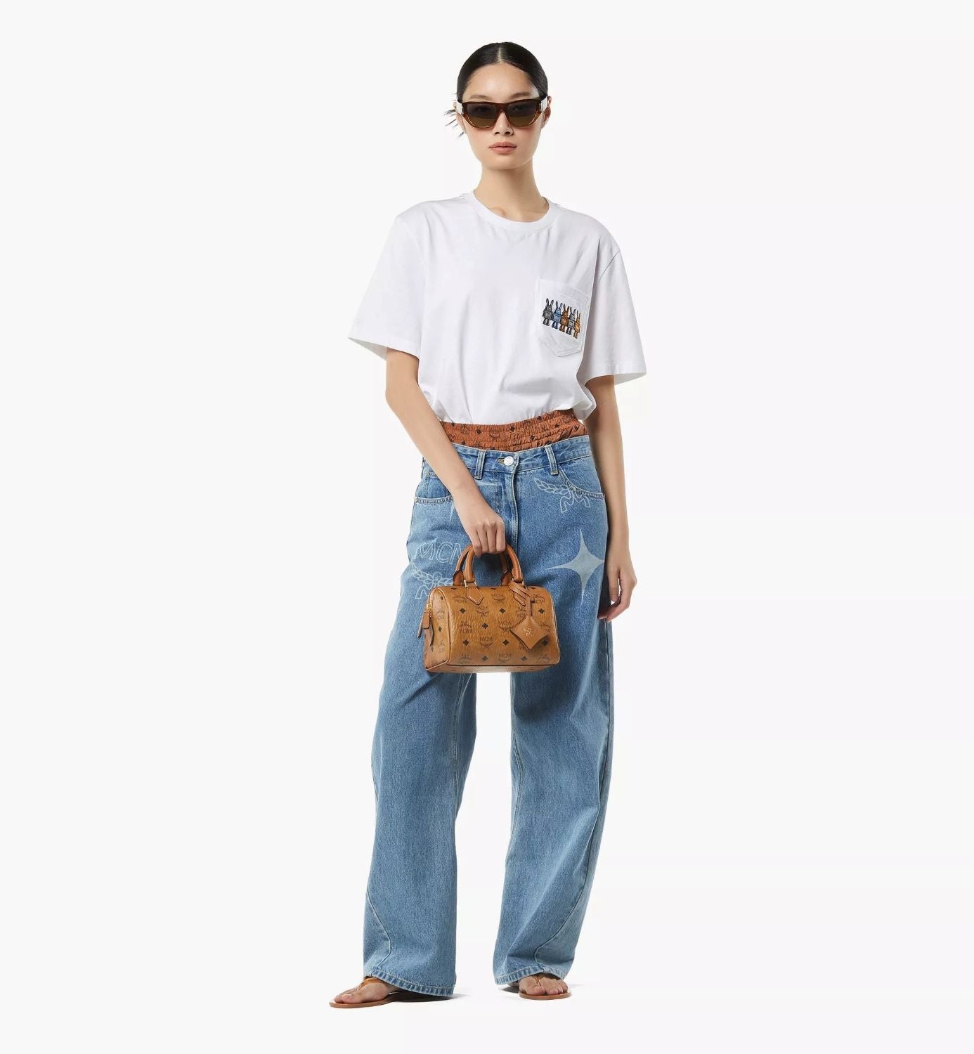 Person wearing a white t-shirt with graphics, blue jeans, and holding a MCM Small Ella Boston Bag in Visetos Cognac MWBFAEA03CO001 on a white background