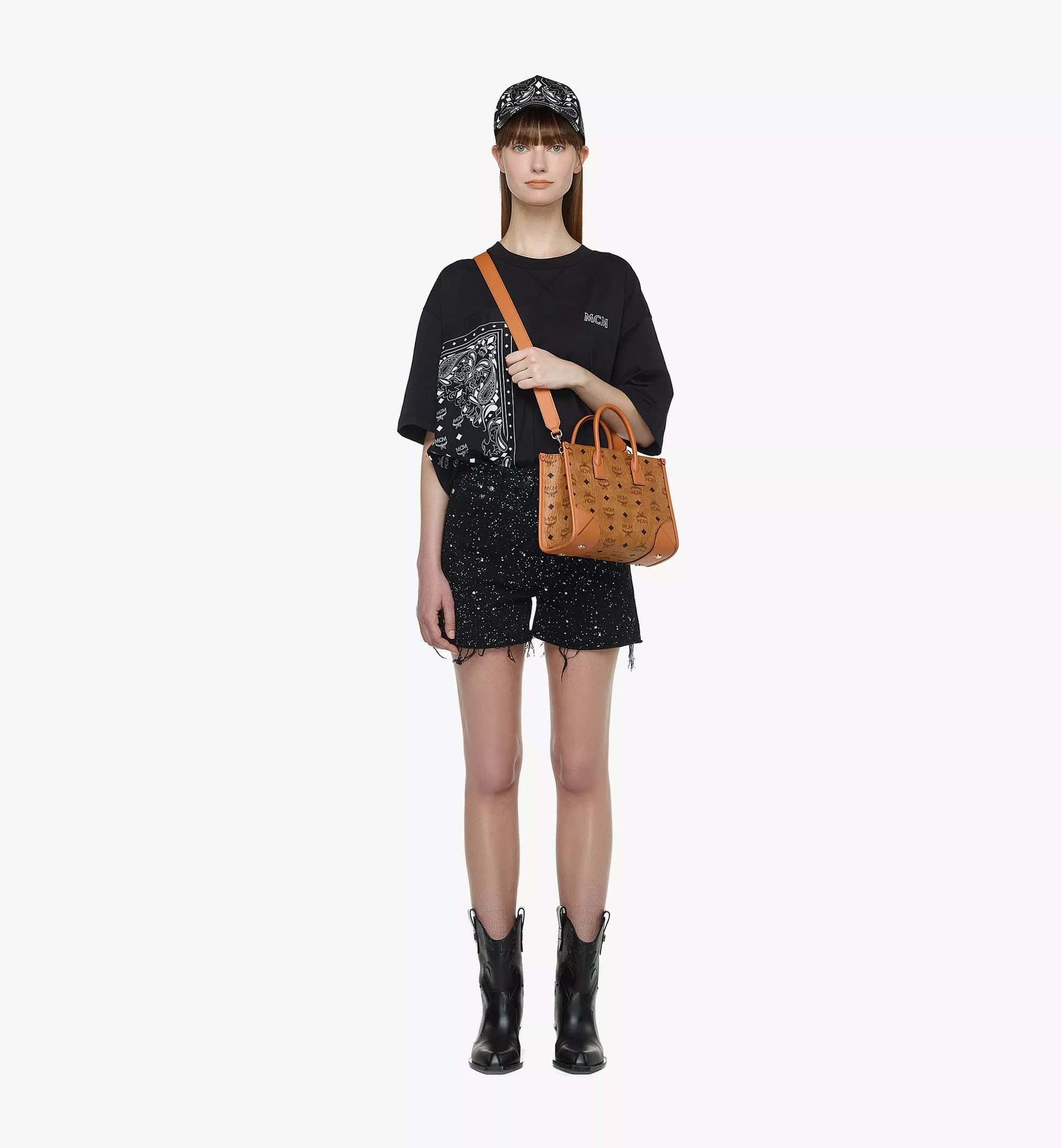 Person wearing a black outfit with a MCM Small München Tote in Visetos Cognac MWTDSBO06CO001 on a white background