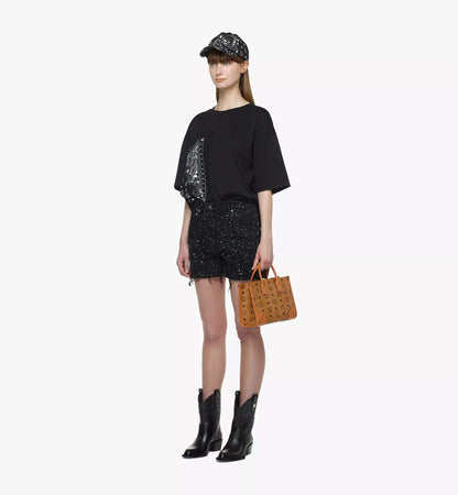 Woman wearing a black sequin top, skirt, and boots with a cap and MCM Small München Tote in Visetos Cognac MWTDSBO06CO001 on a white background