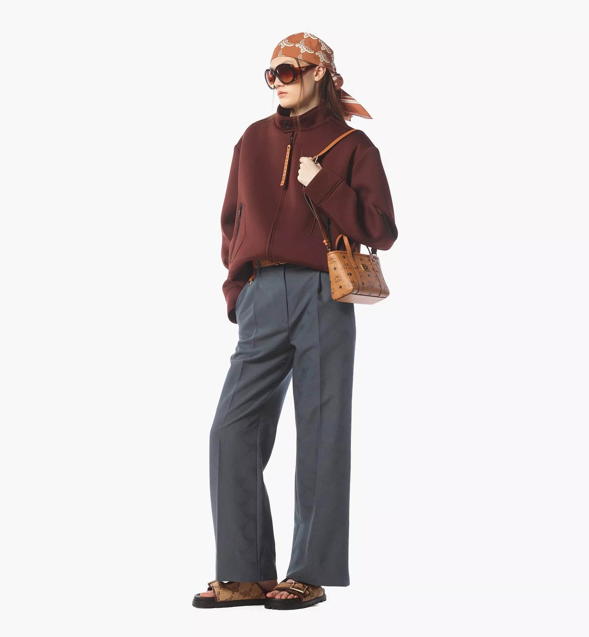 Person wearing a brown sweater, gray pants, and sunglasses, holding a MCM X-Mini Toni Top-Zip Shopper in Visetos Cognac MWPFSMT03CO001 on a white background