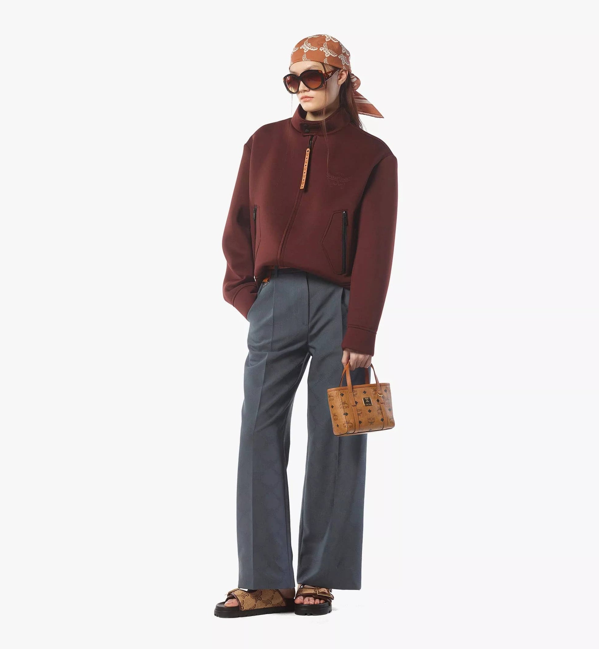 Person wearing a brown jacket, gray pants, and holding a MCM X-Mini Toni Top-Zip Shopper in Visetos Cognac MWPFSMT03CO001 on a white background