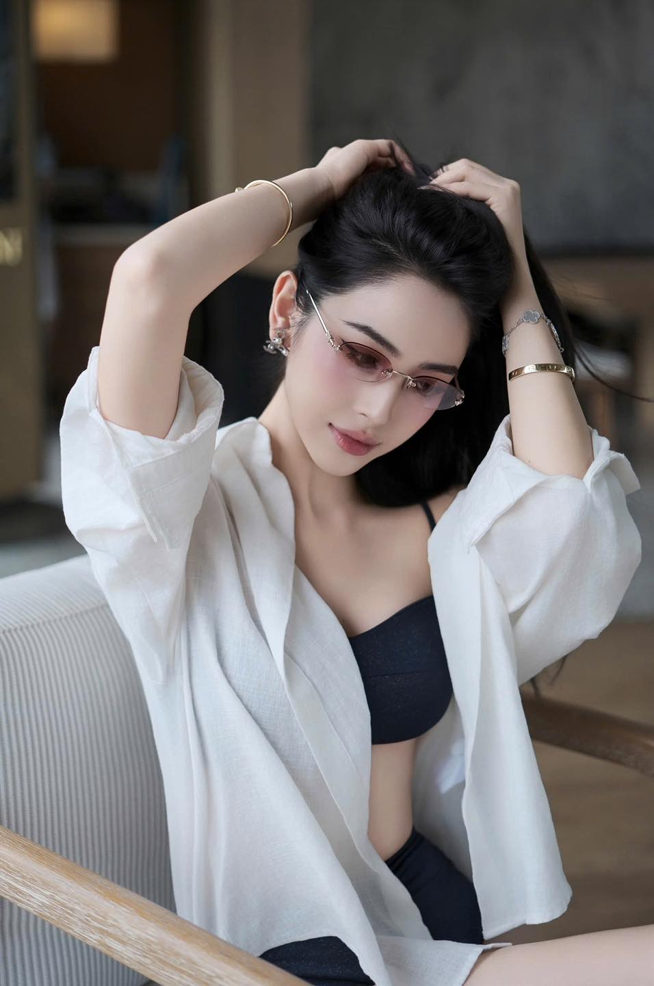 Woman sitting indoors wearing a Miu Miu MU A54S ZVN70O Logo Sunglasses Gradient Pink Lenses SMUA54_EZVN_FE70O_C_051 with a white shirt and black top, adjusting her hair.
