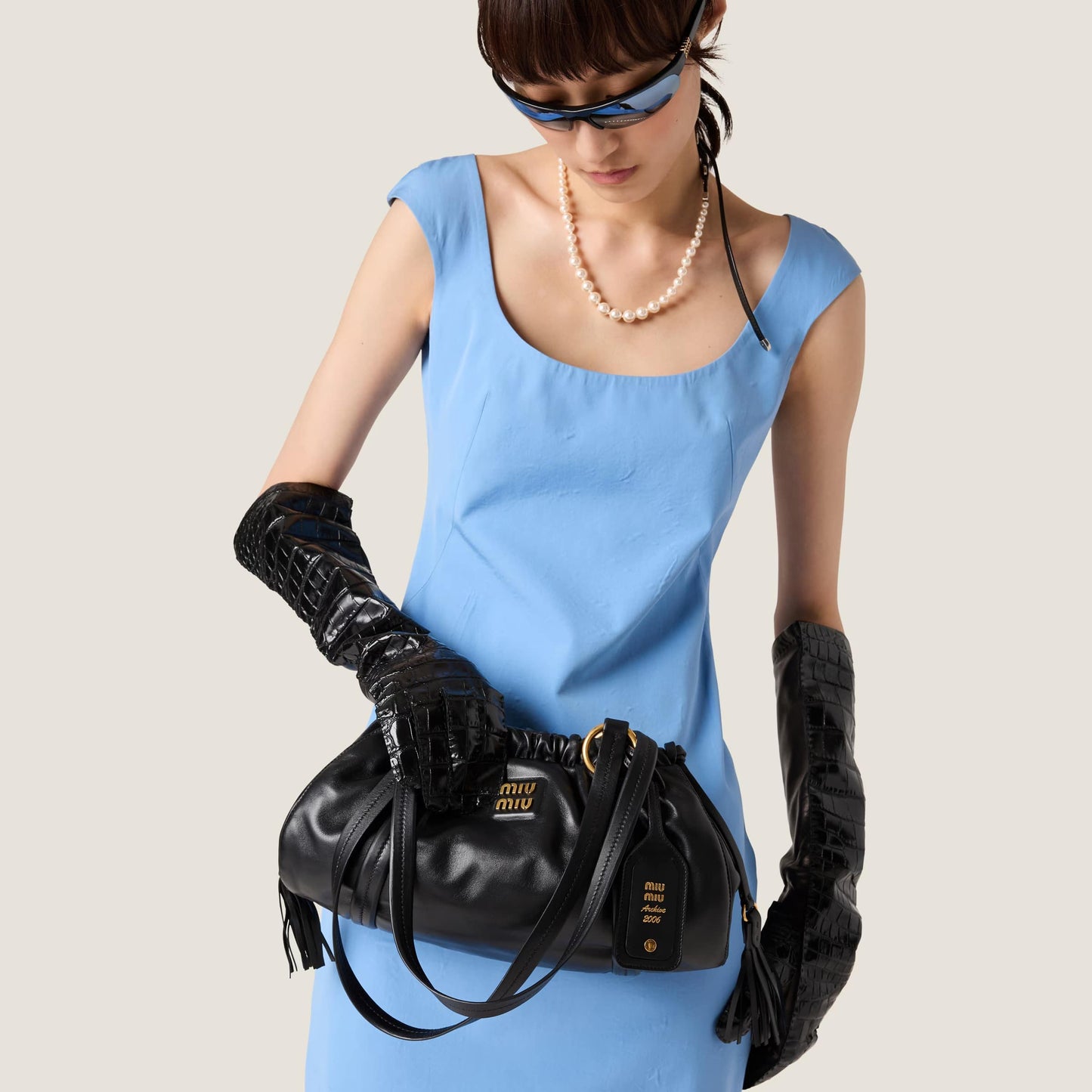 Woman in a blue dress holding a Miu Miu Black Joie Nappa Leather Bag 5BG306_ACSW_F0002_V_OOO with 'Miu Miu' logo, wearing black gloves and sunglasses.