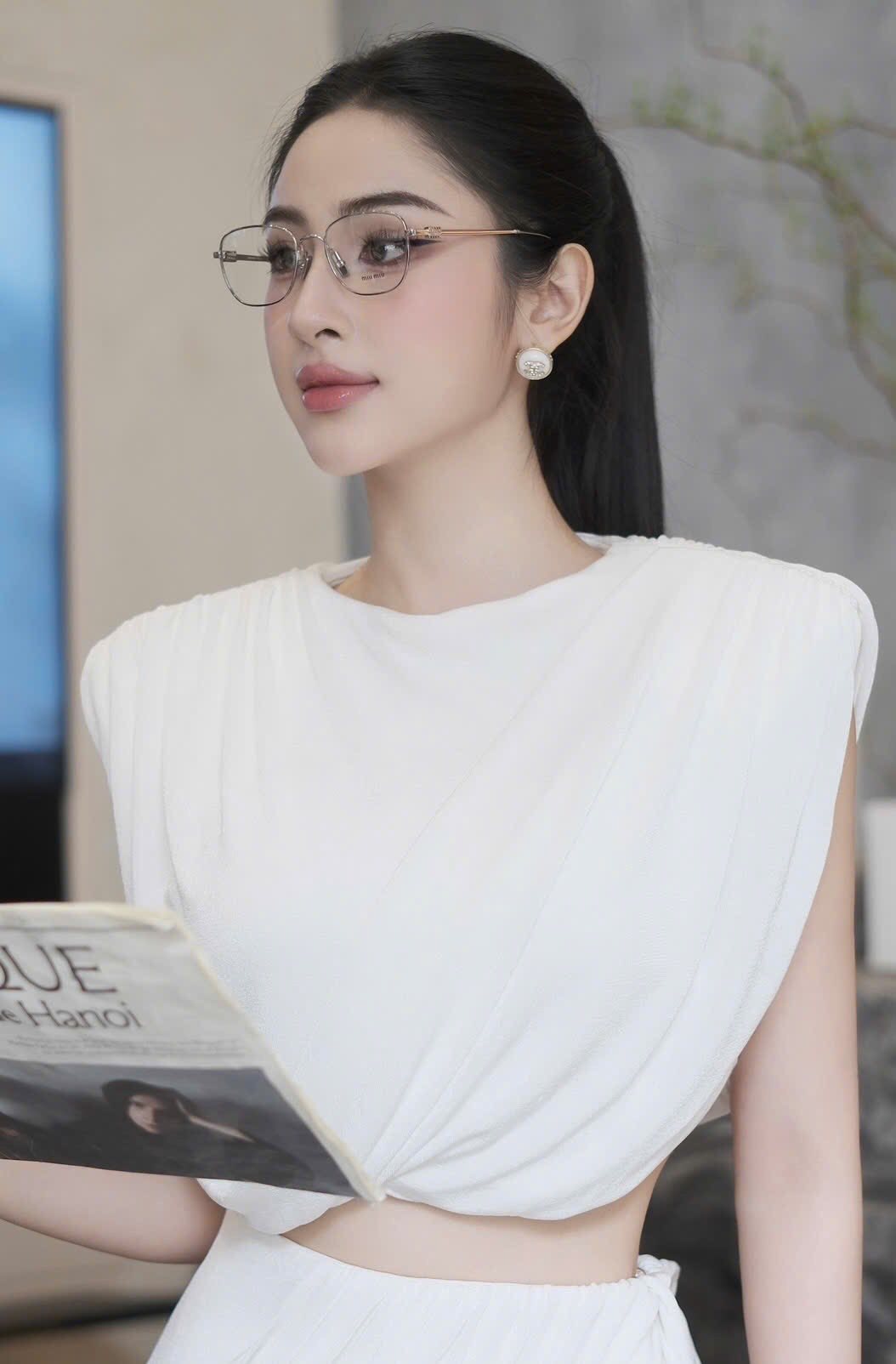 Woman wearing a white top with ruched shoulders and Miu Miu MU 50WV 1BC1O1 Eyeglasses, holding a magazine.