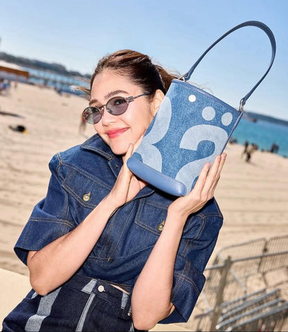 Woman wearing Miu Miu MU 52YS 1BC20R Silver Sunglasses Celeste Lenses SMU52Y_E1BC_FE20R_C_054 holding a blue handbag with abstract patterns on a beach.
