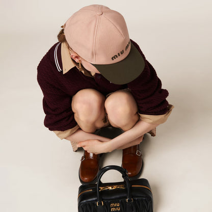 Person wearing a maroon sweater, Miu Miu Orchid Pink Military Drill Baseball Cap 5HC179 ACQ1 F03RG, and brown shoes with a black handbag on a light background