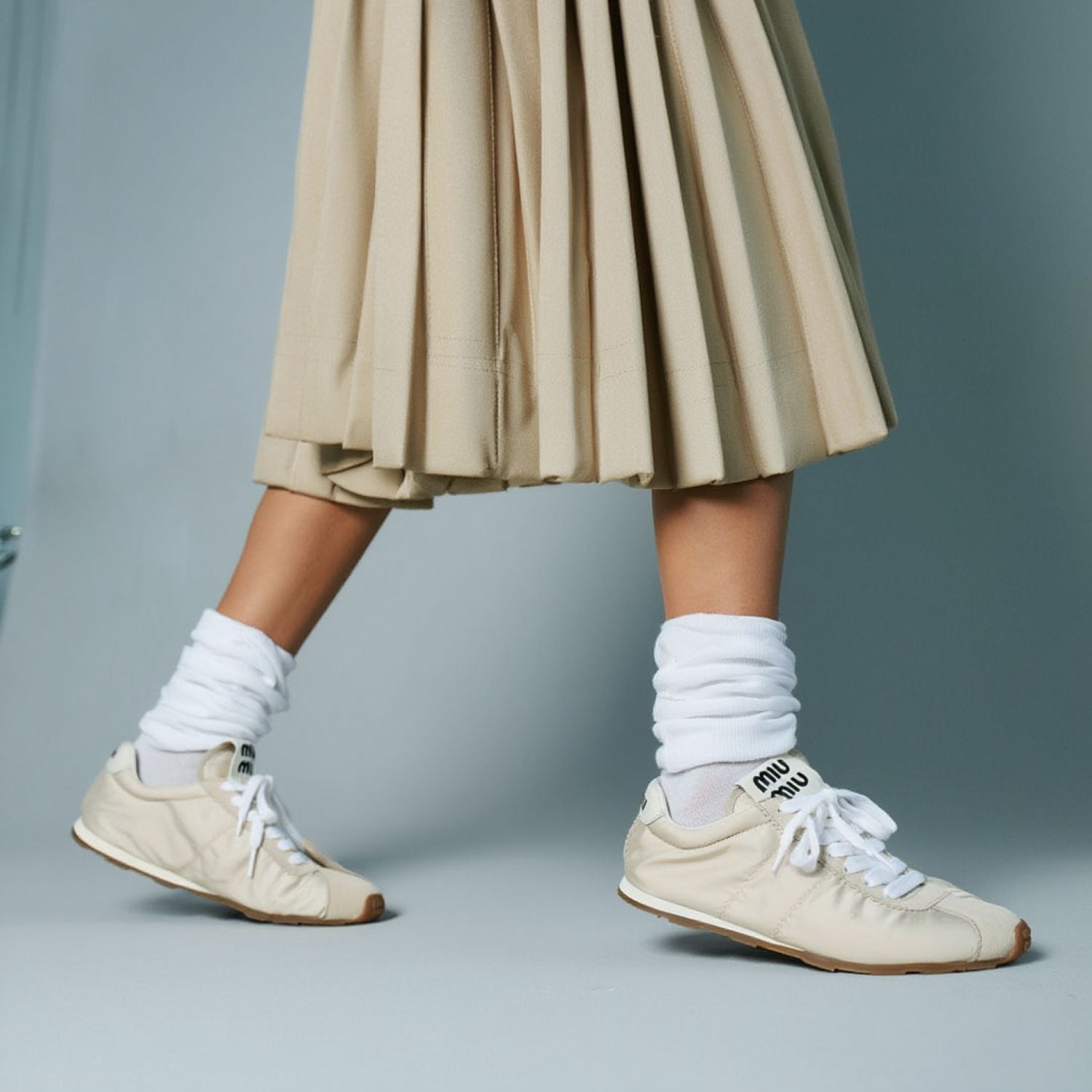 Person wearing Miu Miu Plume Technical Fabric and Suede Sneakers – Ivory 5E114E_D7C_F0304_F_D005, white socks, and a beige pleated skirt on a gray background