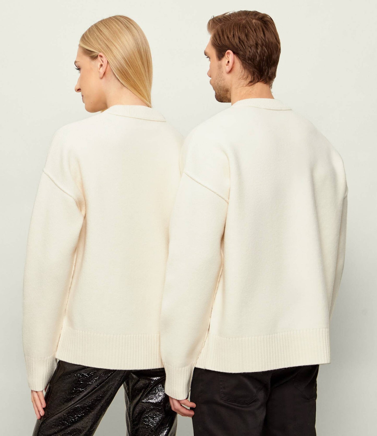 Behind two people wearing Ami Paris Natural Wool Crewneck Sweater With Contrasted Ami De Coeur Off/White UKS838.018.154 and black pants against a plain background