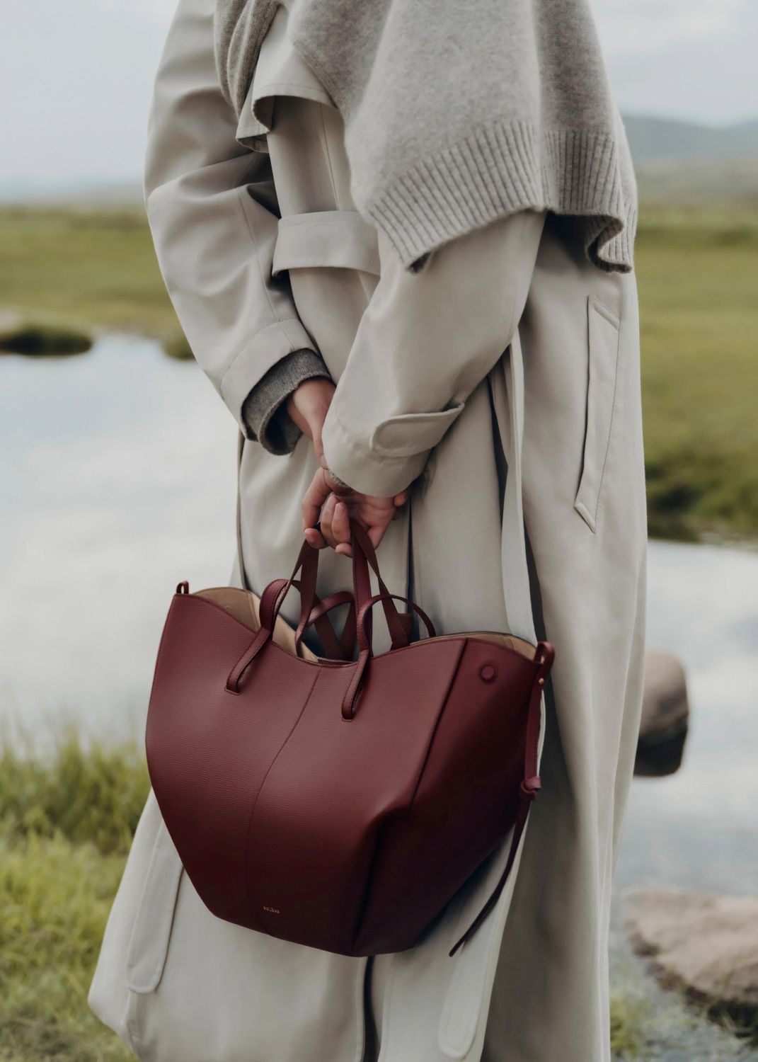 Person holding a mPolène Cyme Edition Textured Black Cherry in a natural setting with water and greenery.