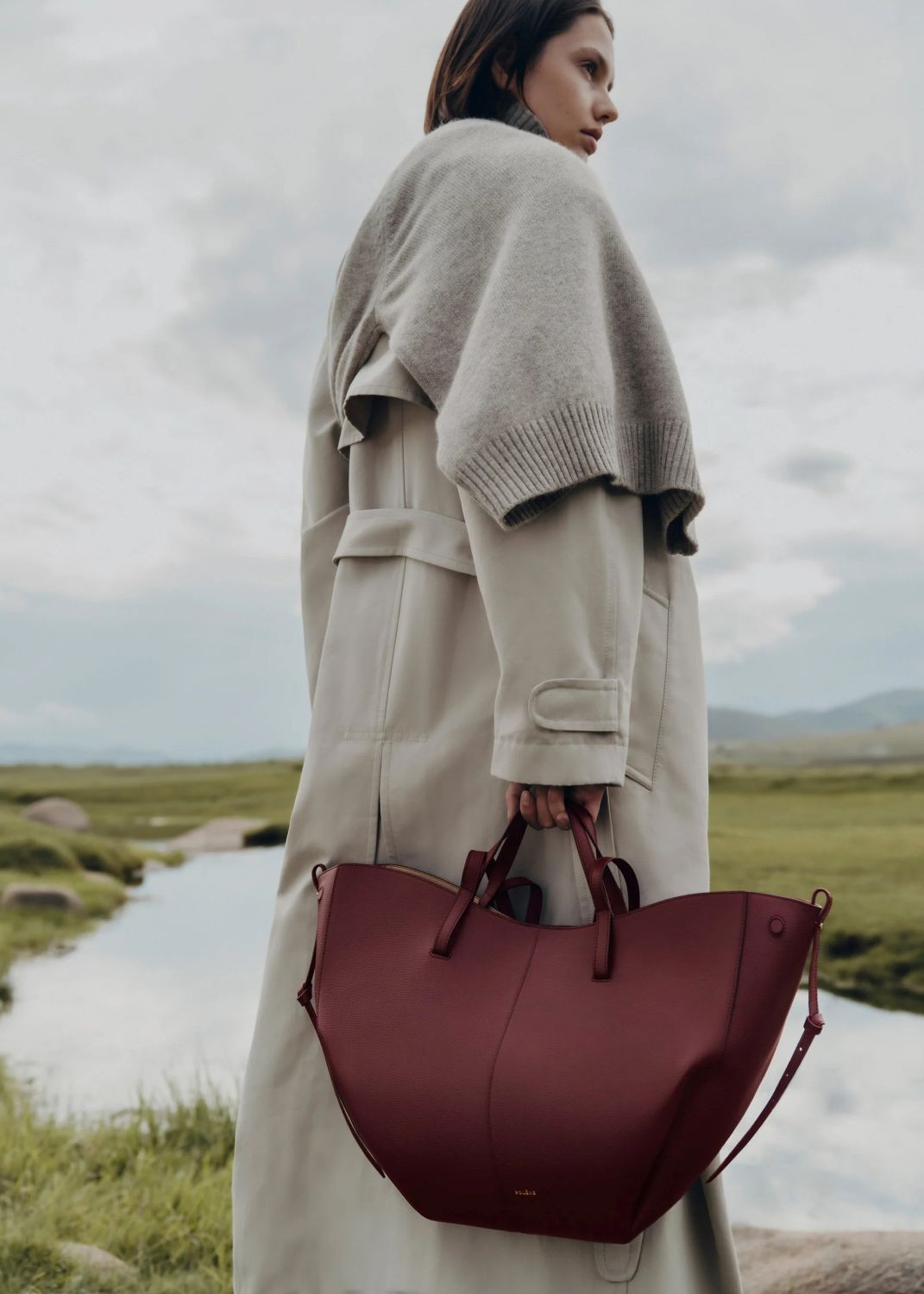 Woman holding a Polène Cyme Edition Textured Black Cherry in a scenic outdoor setting with a lake and mountains.