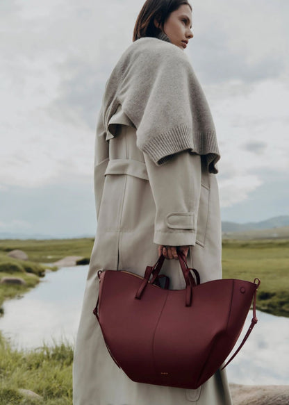Woman holding a Polène Cyme Edition Textured Black Cherry in a scenic outdoor setting with a lake and mountains.