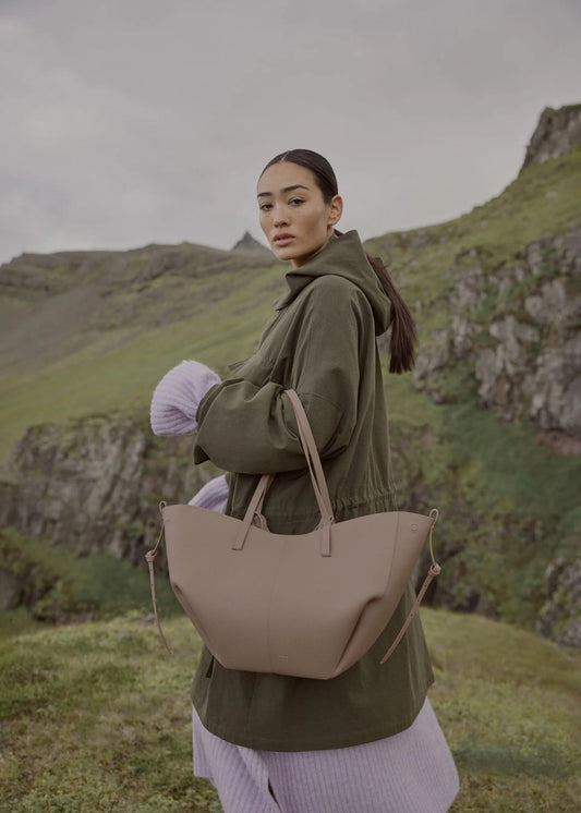 Woman holding a Polène Cyme Edition Textured Taupe in a scenic outdoor setting with green hills and a cloudy sky.