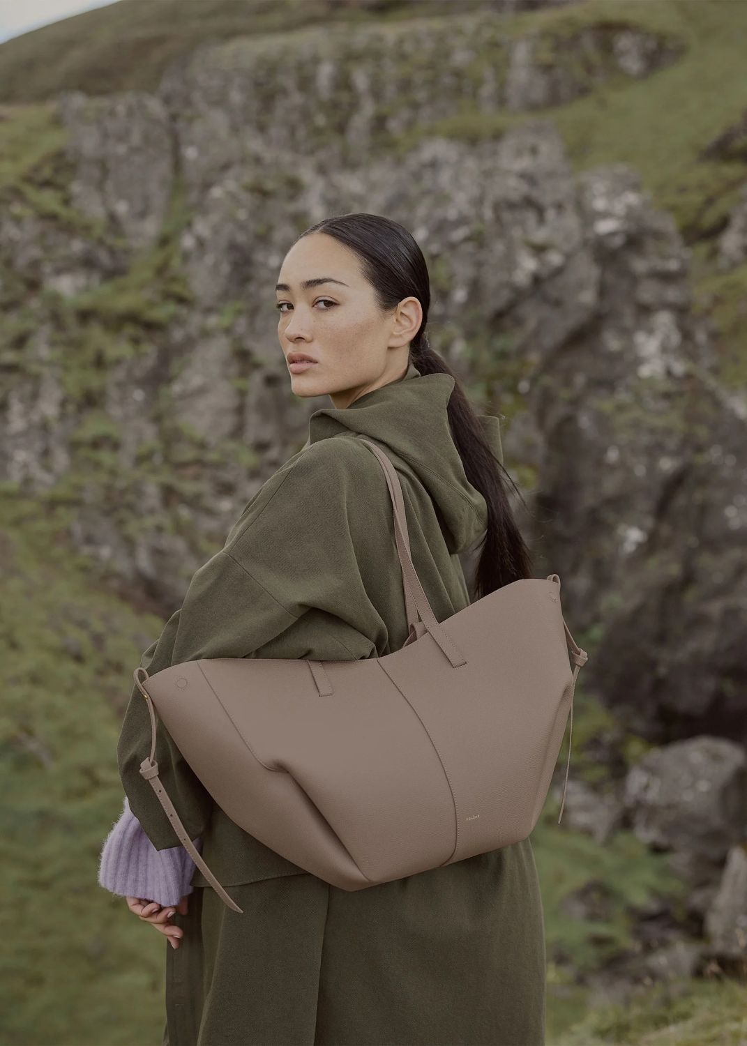 Woman holding a Polène Cyme Edition Textured Taupe in a natural setting with rocky and grassy landscape.