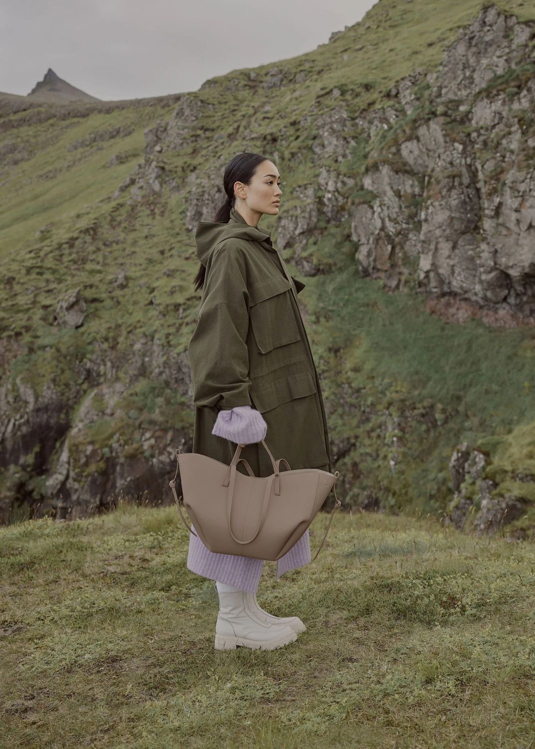 Woman in a green coat holding a Polène Cyme Edition Textured Taupe in a mountainous landscape