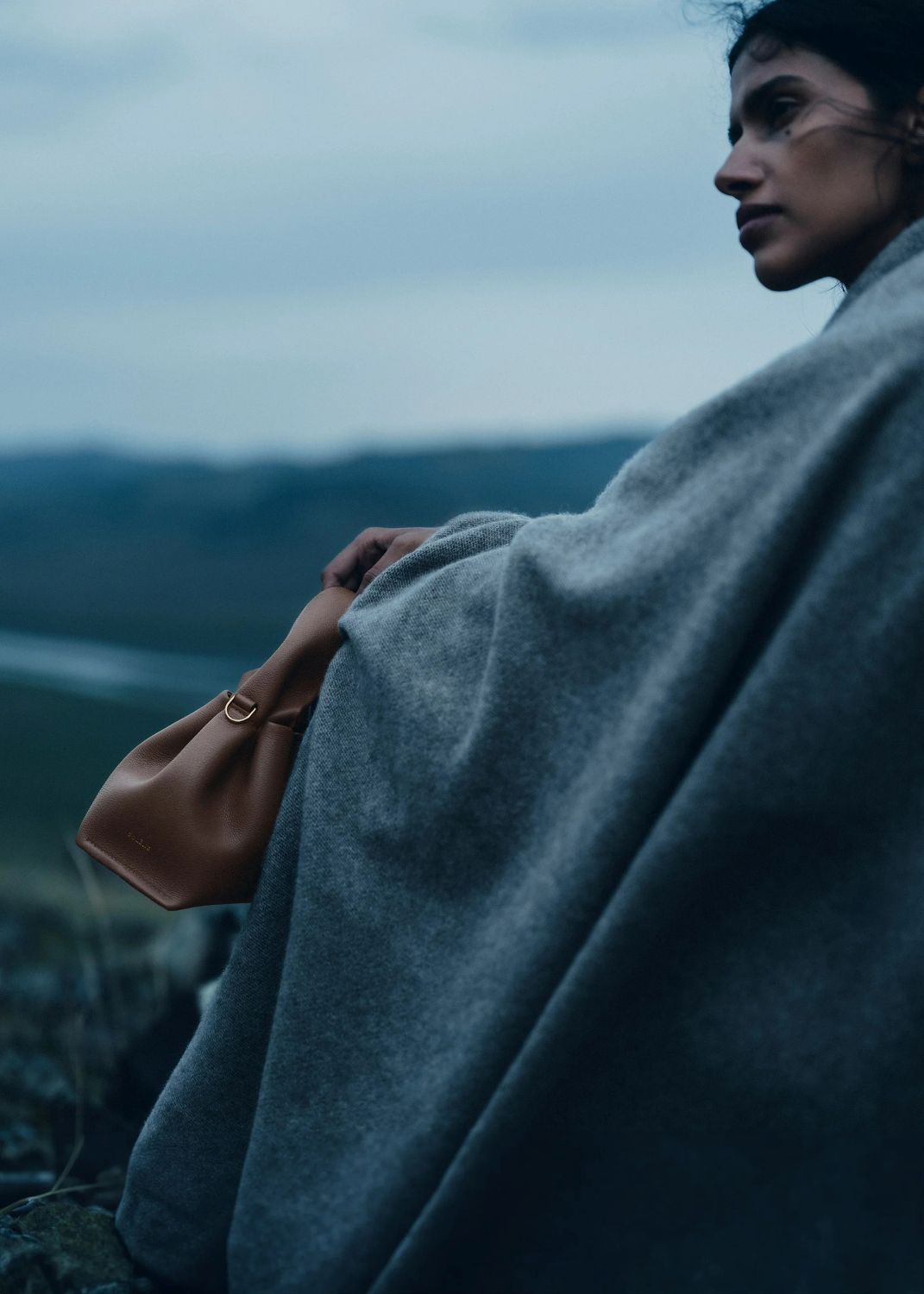 Person in a gray coat holding a Polène Numéro Neuf Mini Edition Textured RootPolène Numéro Neuf Mini Edition Textured Root against a mountainous landscape