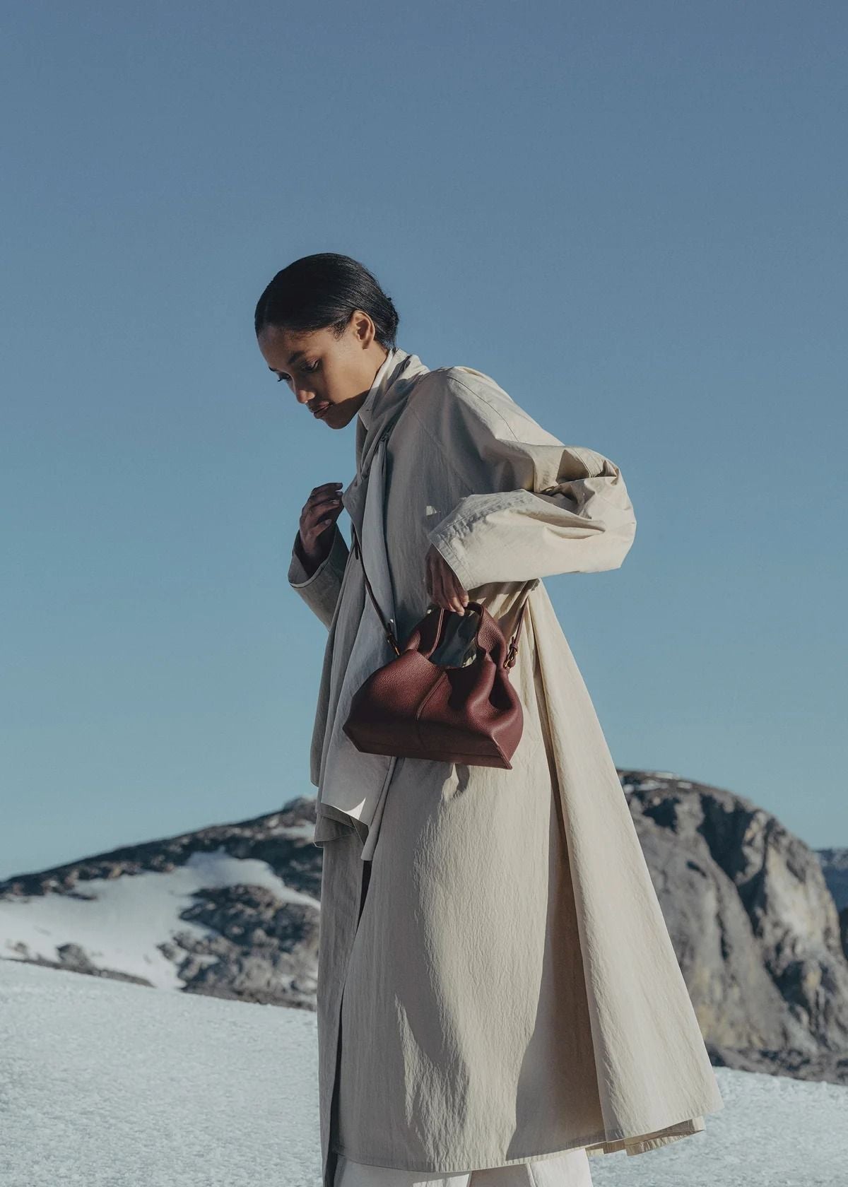 Person wearing a long coat with a Polène Numéro Neuf Mini Edition Textured Black Cherry against a mountainous background