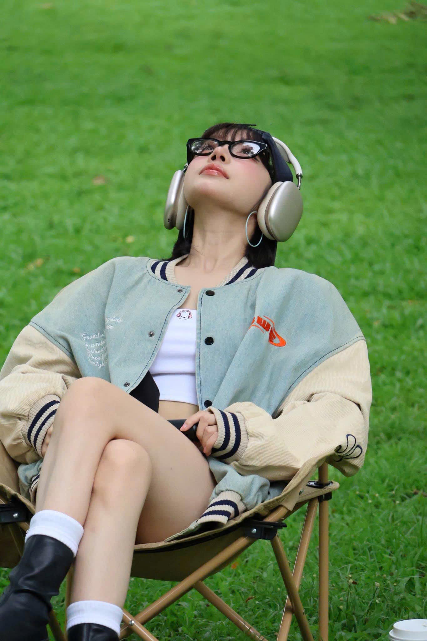 Girl sitting on a chair outdoors with headphones on, wearing a Prada PR A11VD 16K1O1 Oval Black Eyeglasses in light-colored jacket and white sneakers.