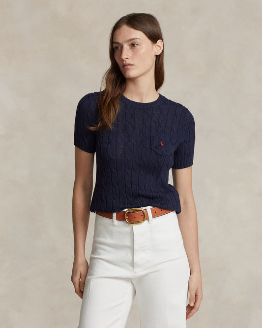 Woman wearing a Polo Ralph Lauren Cable-Knit Cotton Short-Sleeve Jumper – Hunter Navy 34175089504 with a red logo, white pants, and a brown belt against a beige background.