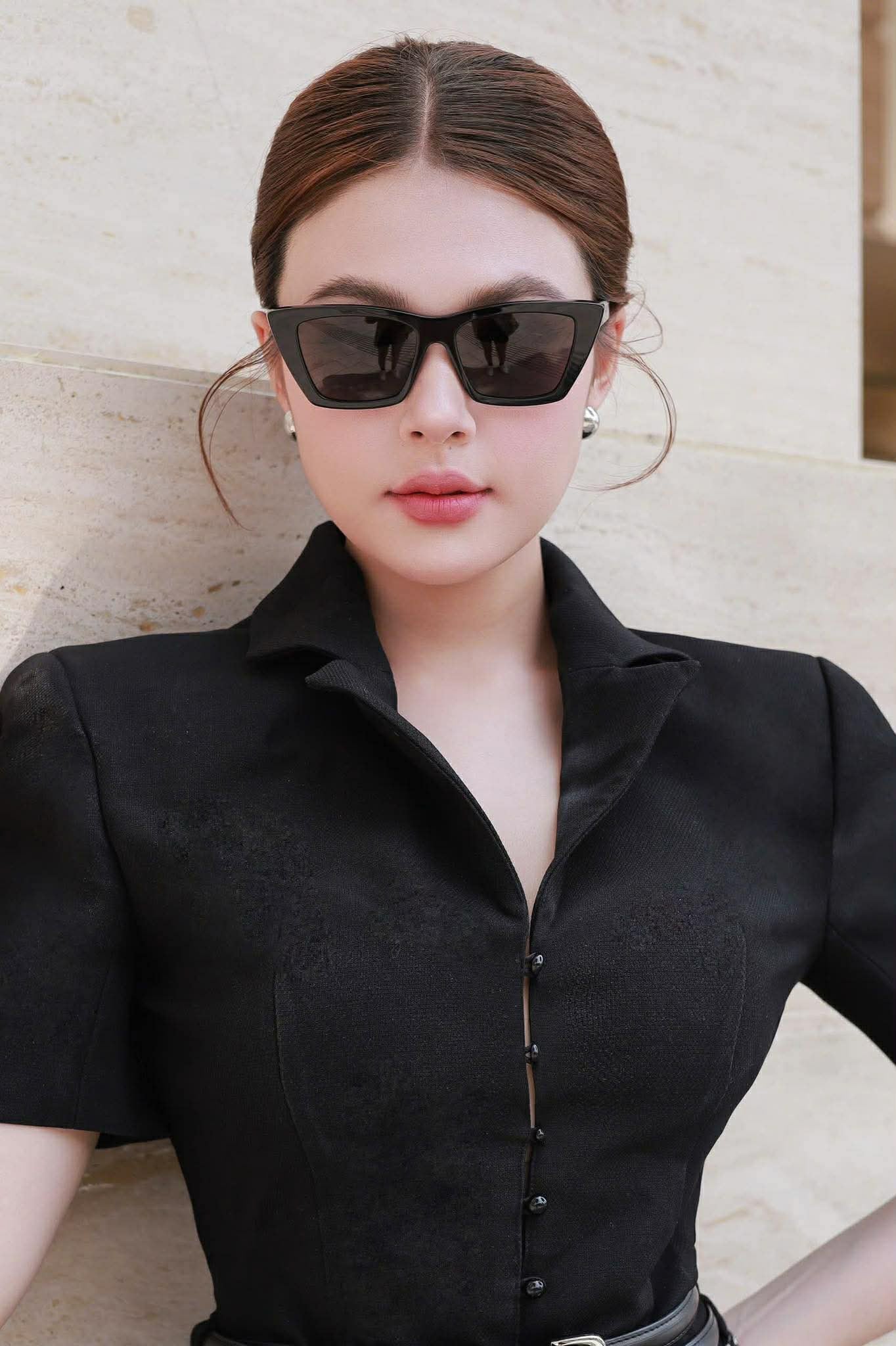 Woman wearing a black shirt and Saint Laurent SL 276 MICA-001 Square Cat-Eye Sunglasses against a light-colored wall.