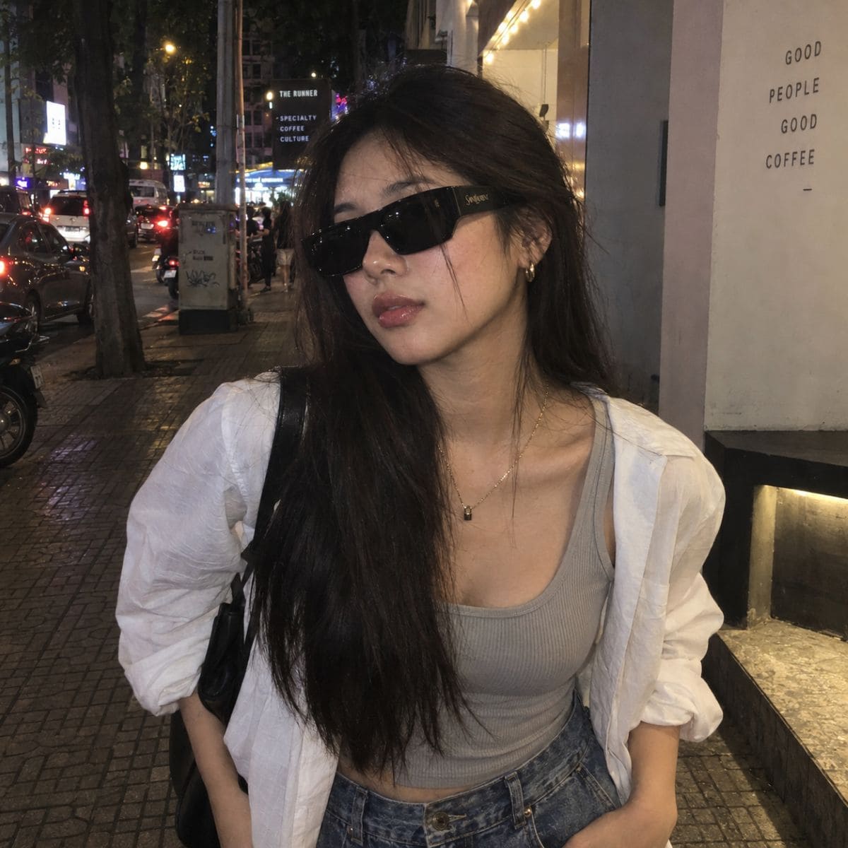 A woman wearing Saint Laurent SL 740 005, a white button-up shirt, a gray ribbed tank top, and blue jeans, standing on a city sidewalk at night.