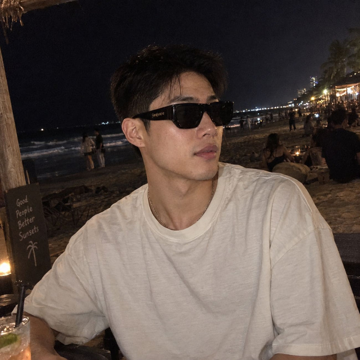 A man wearing Saint Laurent SL 740 005 and a light beige t-shirt sitting at a beach bar at night.