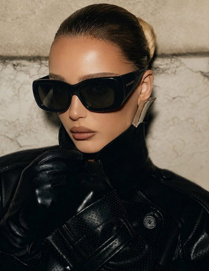 A close-up portrait of a model wearing oversized Saint Laurent SL 901 HOWL-001, a black leather jacket, and a black leather glove, set against a textured beige stone wall.