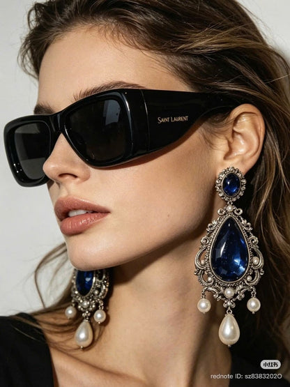 A close-up of a woman wearing black Saint Laurent SL 901 HOWL-001 sunglasses and ornate silver drop earrings with deep blue stones and white pearls, against a light gray background.