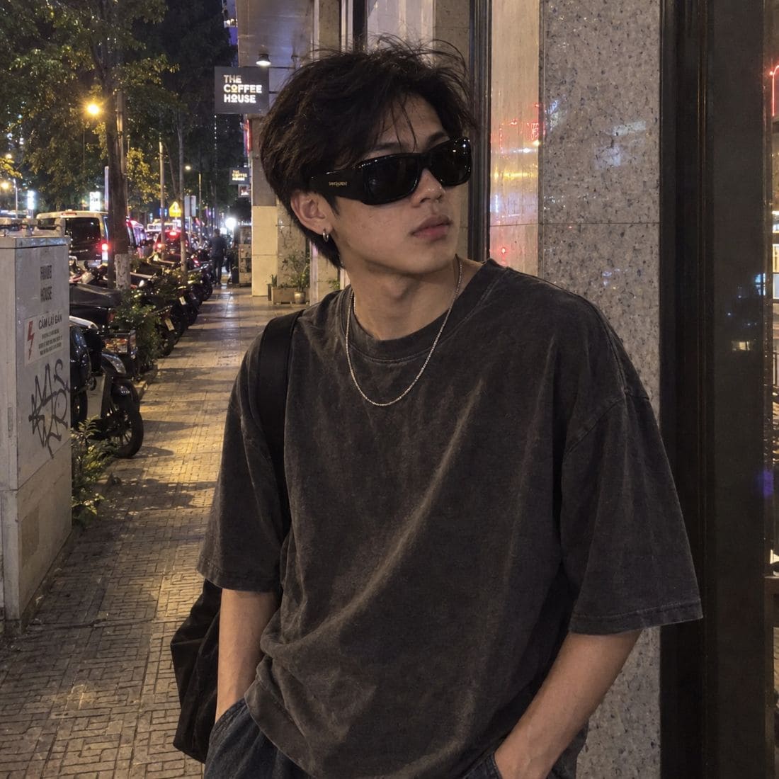 A man wearing a dark washed black oversized t-shirt, Saint Laurent SL 901 HOWL-001, and a silver chain necklace, standing on a city sidewalk at night.