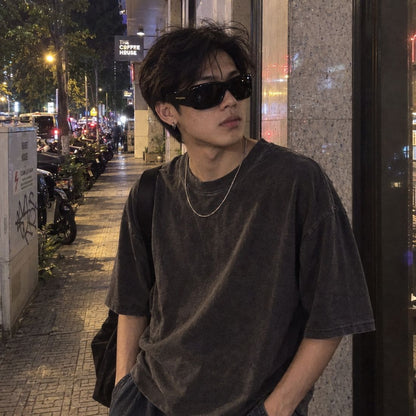 A man wearing a dark washed black oversized t-shirt, Saint Laurent SL 901 HOWL-001, and a silver chain necklace, standing on a city sidewalk at night.