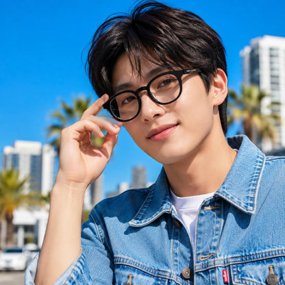 A young man with dark hair wearing Saint Laurent SL 907/F 001 Black Round Eyeglasses, a light blue denim jacket, and a white t-shirt, posing outdoors against a bright blue sky with palm trees and buildings in the background.