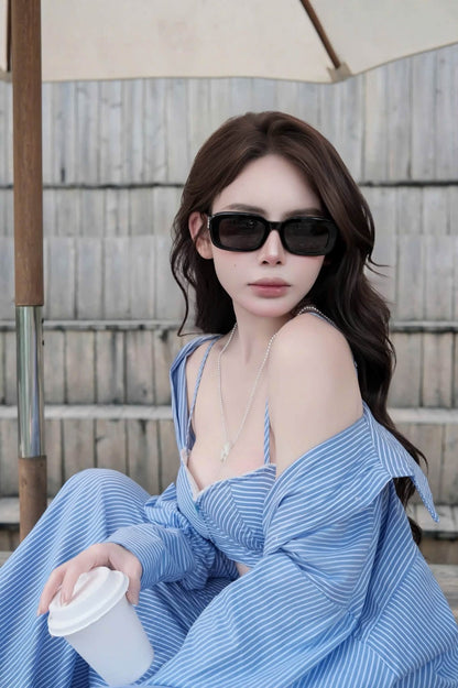 A woman wearing a Saint Laurent SL M130 Black Square Sunglasses in blue and white striped off-the-shoulder dress and matching striped cardigan, black sunglasses, and a delicate silver necklace, holding a white takeaway coffee cup in front of a wooden fence background.