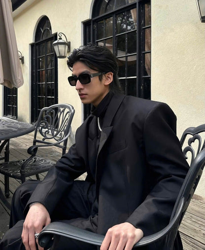 A man wearing a black oversized blazer, black high-neck shirt, and Saint Laurent SL M130 Black Square Sunglasses sits in a black metal patio chair on an outdoor deck in front of a beige stucco building.