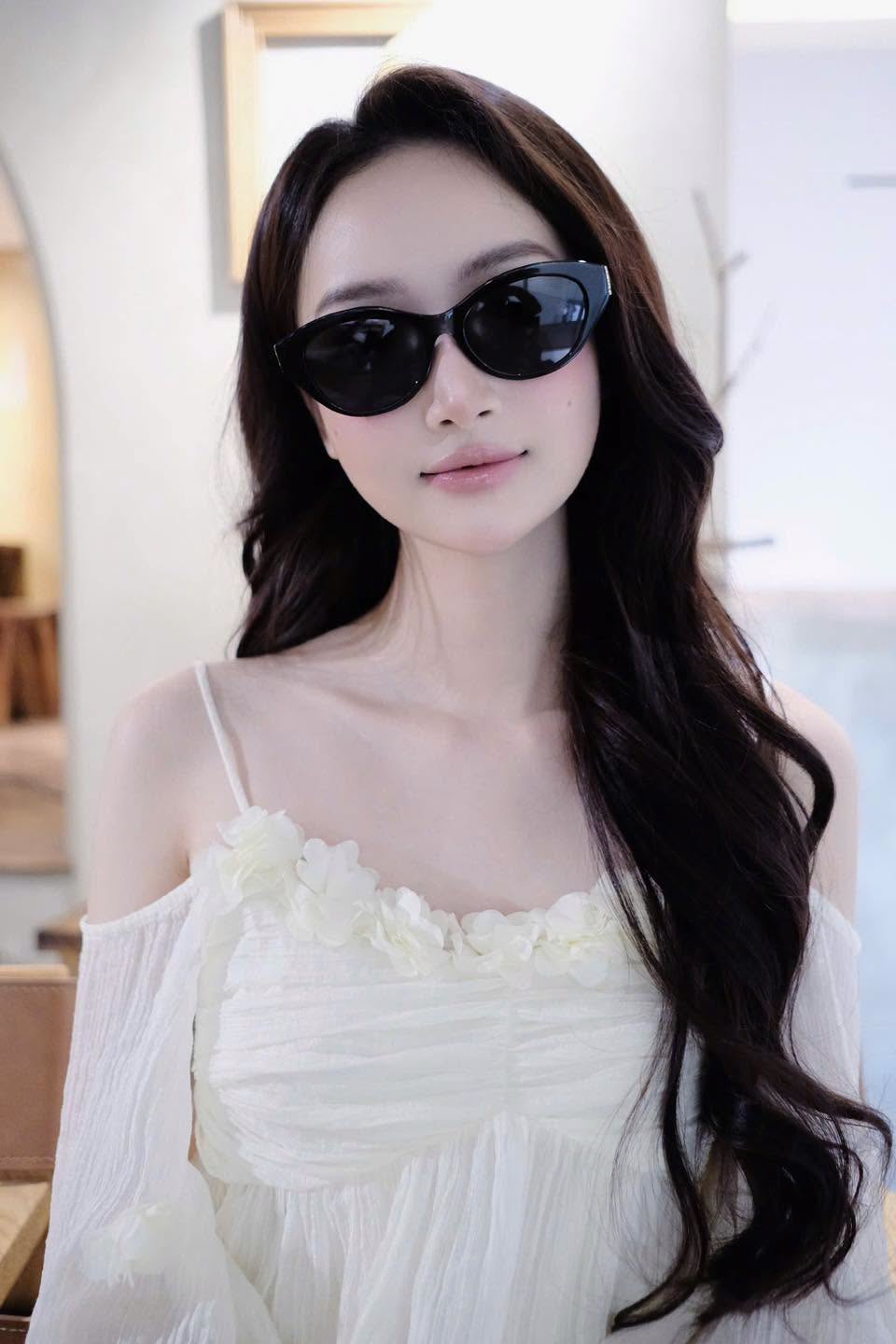 A woman with long wavy dark hair wearing Saint Laurent SL M148 001 Black Round Sunglasses and a white off-the-shoulder dress with floral details, photographed indoors against a softly blurred light background.