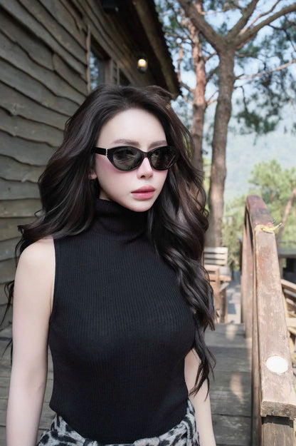 A woman with long wavy dark hair wearing a sleeveless black ribbed turtleneck top and Saint Laurent SL M148 001 Black Round Sunglasses, standing outdoors on a wooden walkway next to a rustic wooden building.