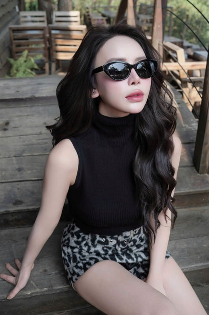A woman with long wavy black hair sits on rustic wooden steps outdoors, wearing Saint Laurent SL M148 001 Black Round Sunglasses, a black sleeveless mock-neck top, and black-and-white patterned shorts.